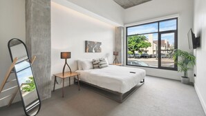 Een moderne slaapkamer met een groot raam, een bed, nachtkastjes, een spiegel en een plant in pot.