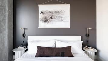 Une chambre à coucher moderne avec un lit blanc, un oreiller marron foncé et une œuvre d’art abstraite accrochée au mur.
