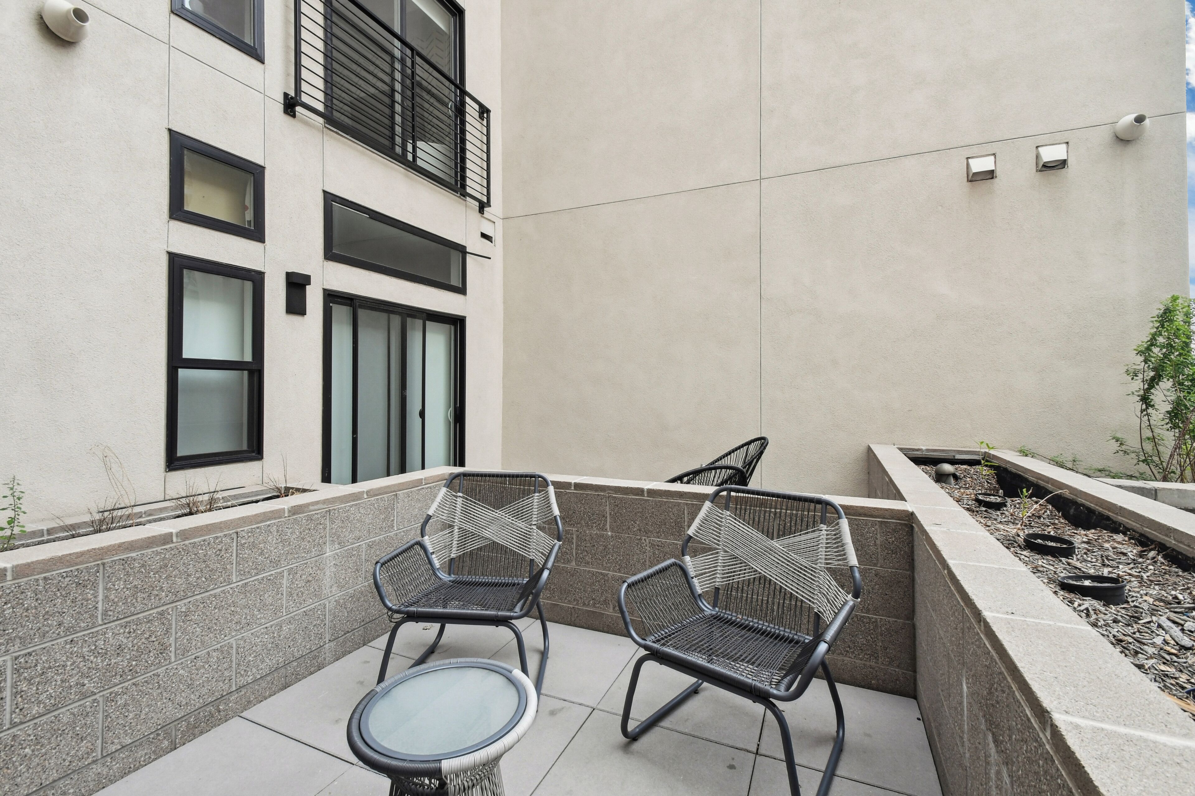 Un patio al aire libre con dos sillas de metal y una mesa de vidrio, un muro bajo con una jardinera, y un edificio con varias ventanas y una puerta.