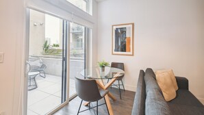 A modern living room with a glass dining table, grey sofa, and a large window offering a view of the city.