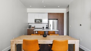 A modern kitchen with white cabinets, a stainless steel refrigerator, and a wooden dining table with orange chairs. - Art Inspired Stay | 1BR LoHi Loft | Dog-Friendly (Denver)