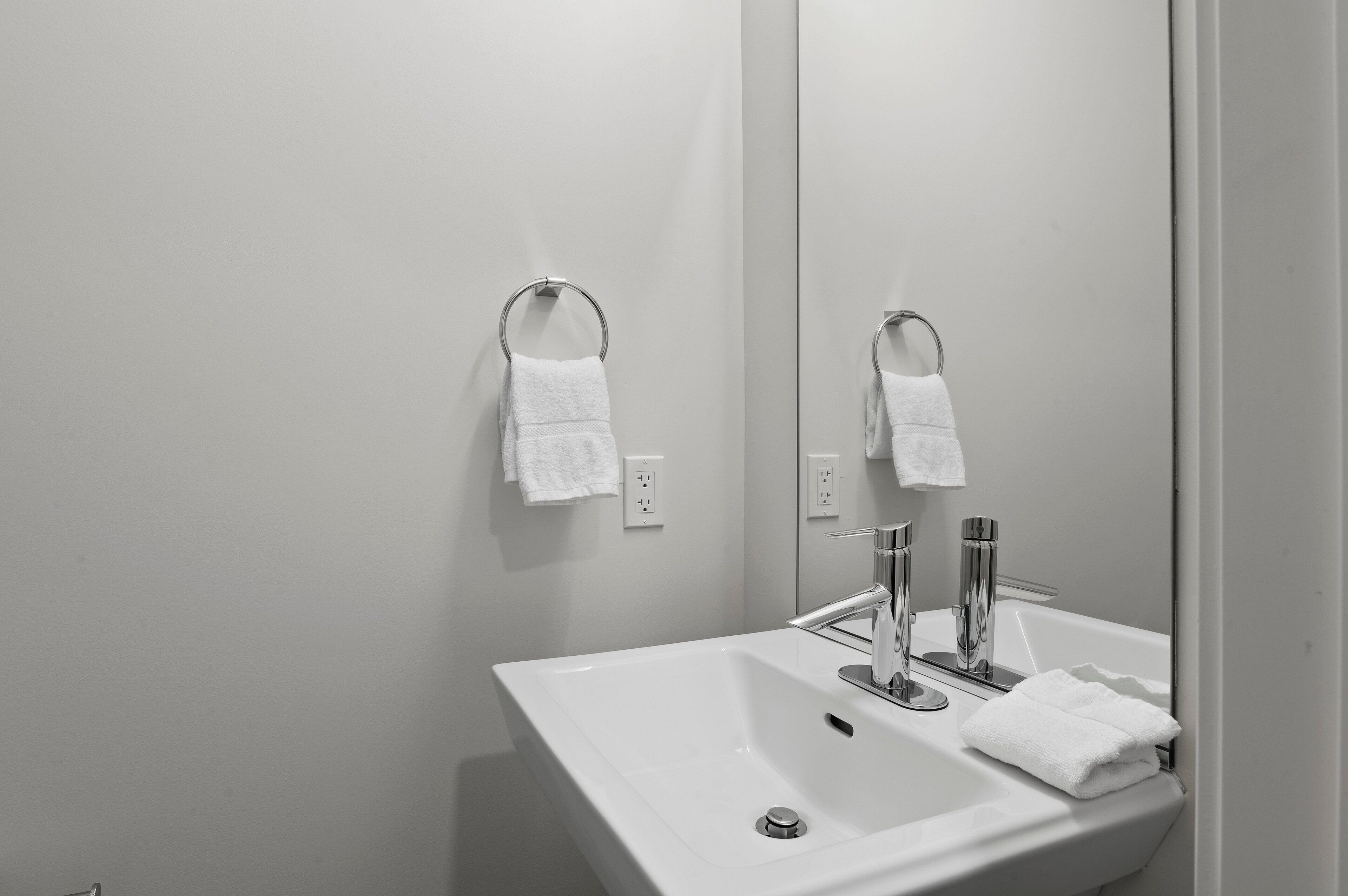 Une salle de bain moderne avec un lavabo blanc, un robinet chromé et un grand miroir. Deux porte-serviettes sont fixés au mur, et une serviette blanche est suspendue à chacun d’eux.