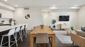 Une cuisine moderne avec des meubles blancs, une table à manger en bois et des tabourets de bar. Un téléviseur est fixé au mur et une plante décore la table à manger.