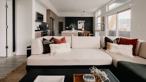A modern living room with a large sectional sofa, a dining area with a kitchen in the background, and large windows providing natural light. - Rhythm of Night | 2BR LoHi Loft w/ Balcony (Denver)