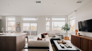 A modern living room with a sofa, coffee table, and TV. There is a kitchen area with a sink and cabinets. Large windows provide natural light and views of the outside. - Rhythm of Night | 2BR LoHi Loft w/ Balcony (Denver)