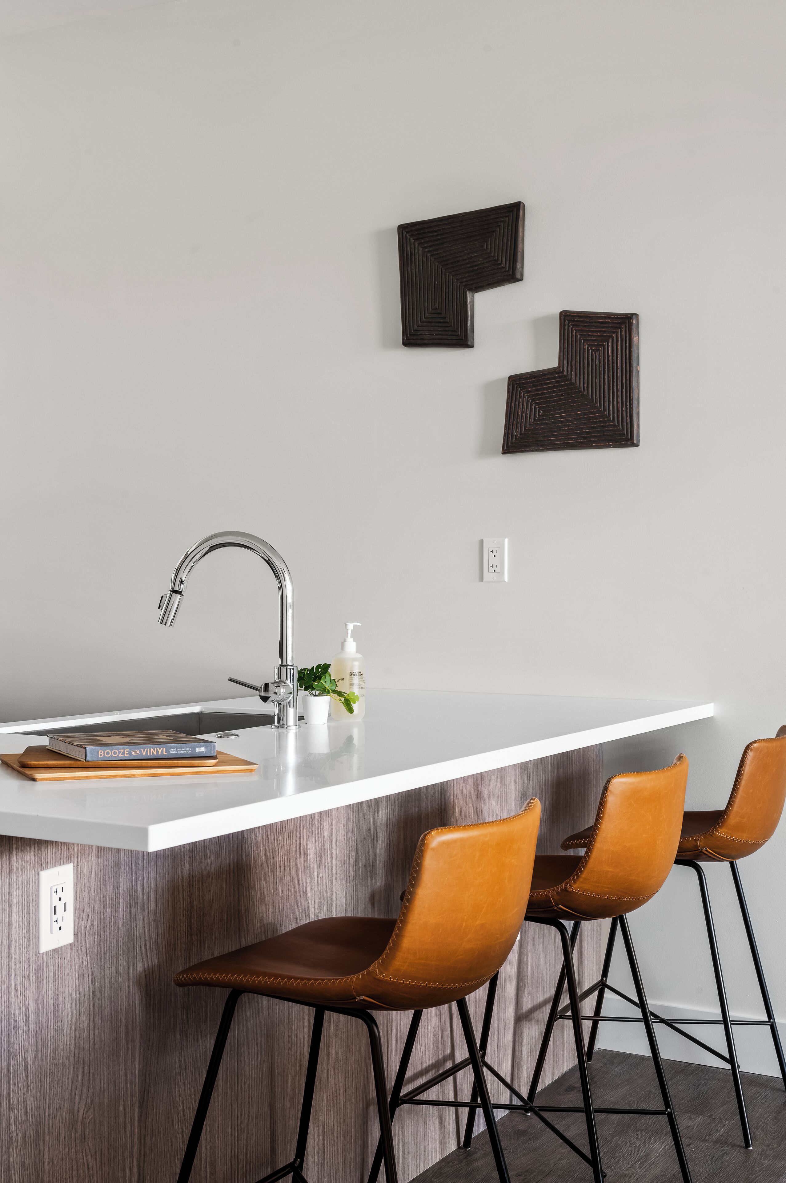 A modern kitchen with a white countertop, a stainless steel sink, and three brown leather bar stools.