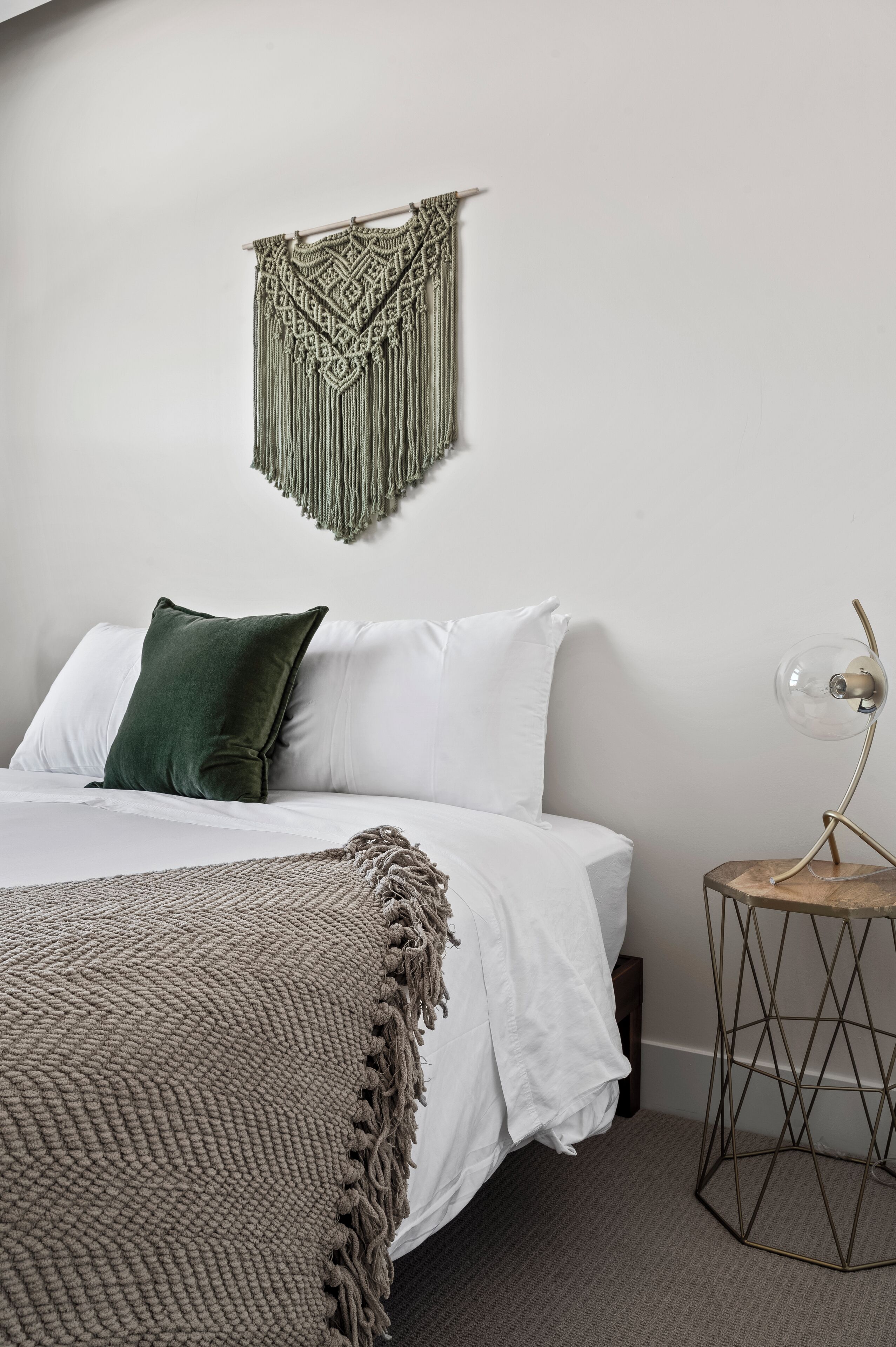 Ein ordentlich bezogenes Bett mit weißen Leinen, einem grünen Kopfkissen und einer strukturierten Tagesdecke. Darüber hängt ein Wandbehang aus Makramee, und ein kleiner runder Tisch mit einer Lampe steht daneben.