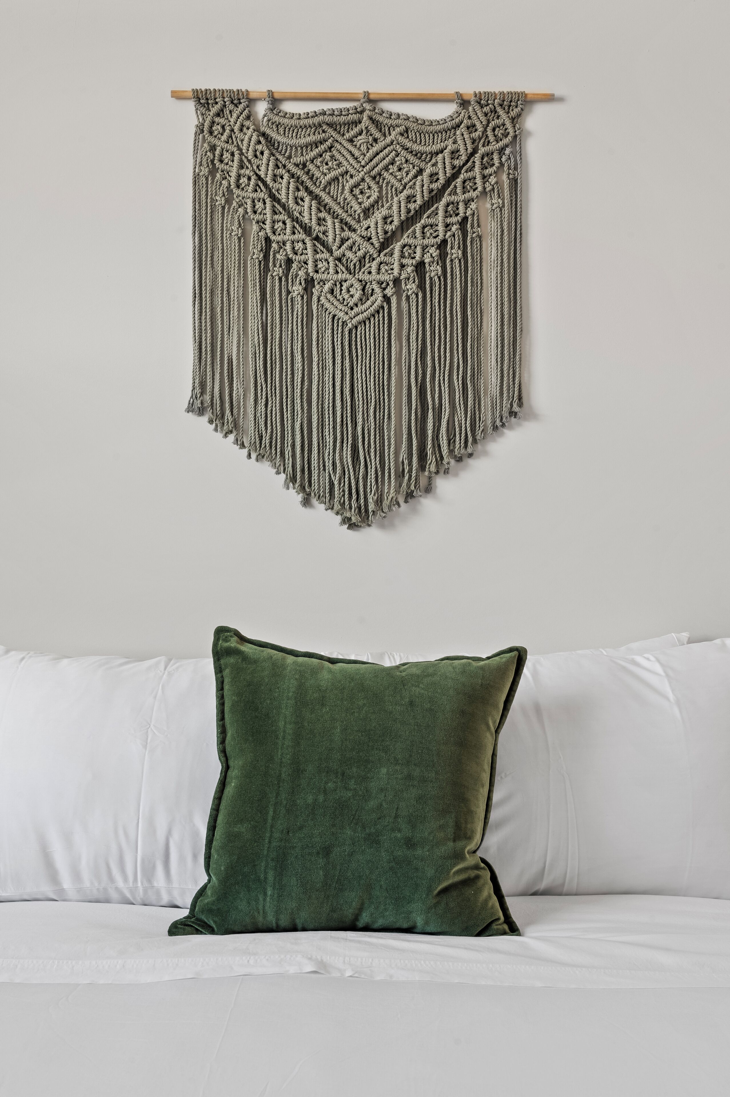 Een macramé wanddecoratie met een houten lat, een groen kussen op een witte bank, en een wit kussen.