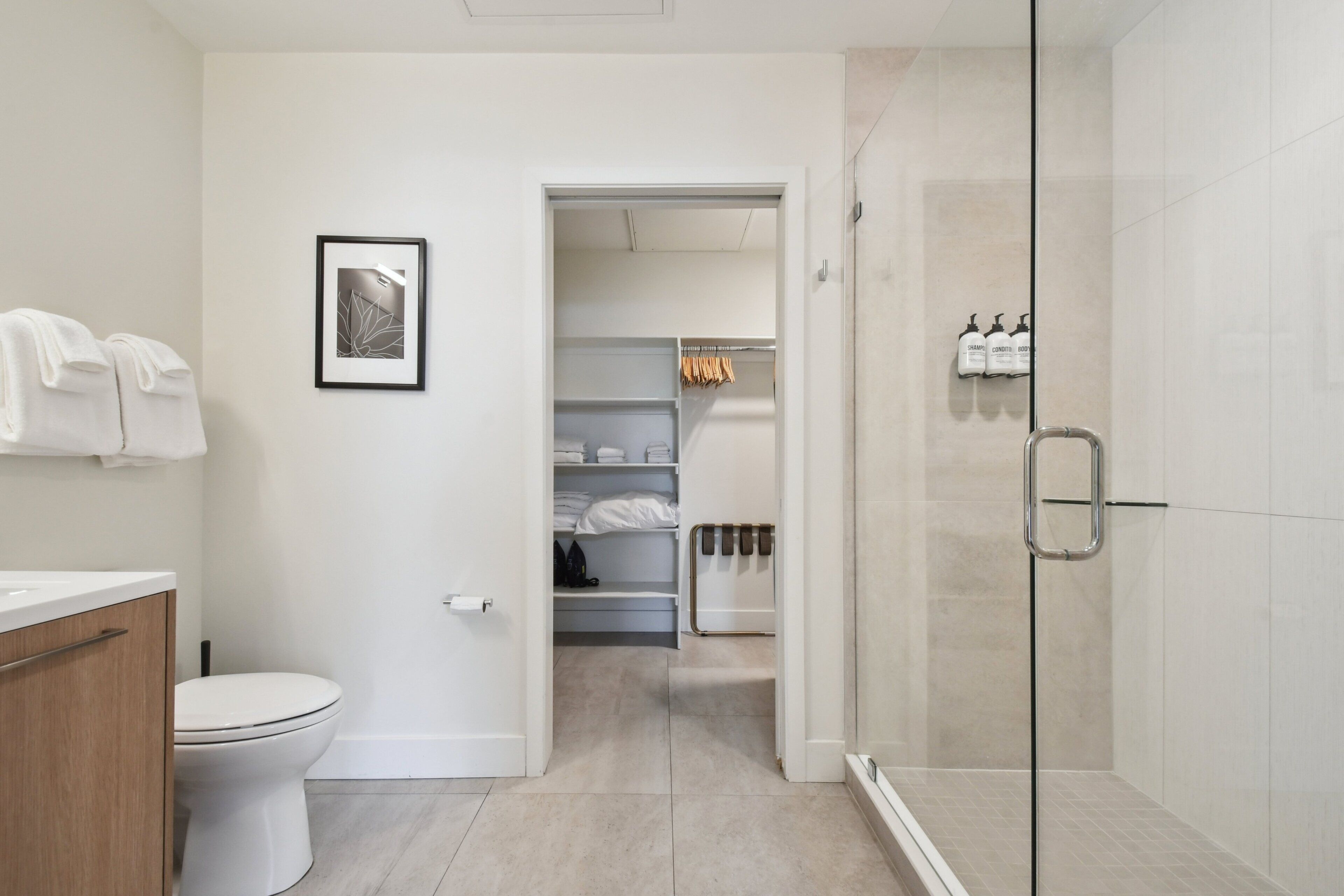 A modern bathroom with a toilet, a shower with glass doors, and a walk-in closet with shelves and hanging clothes.