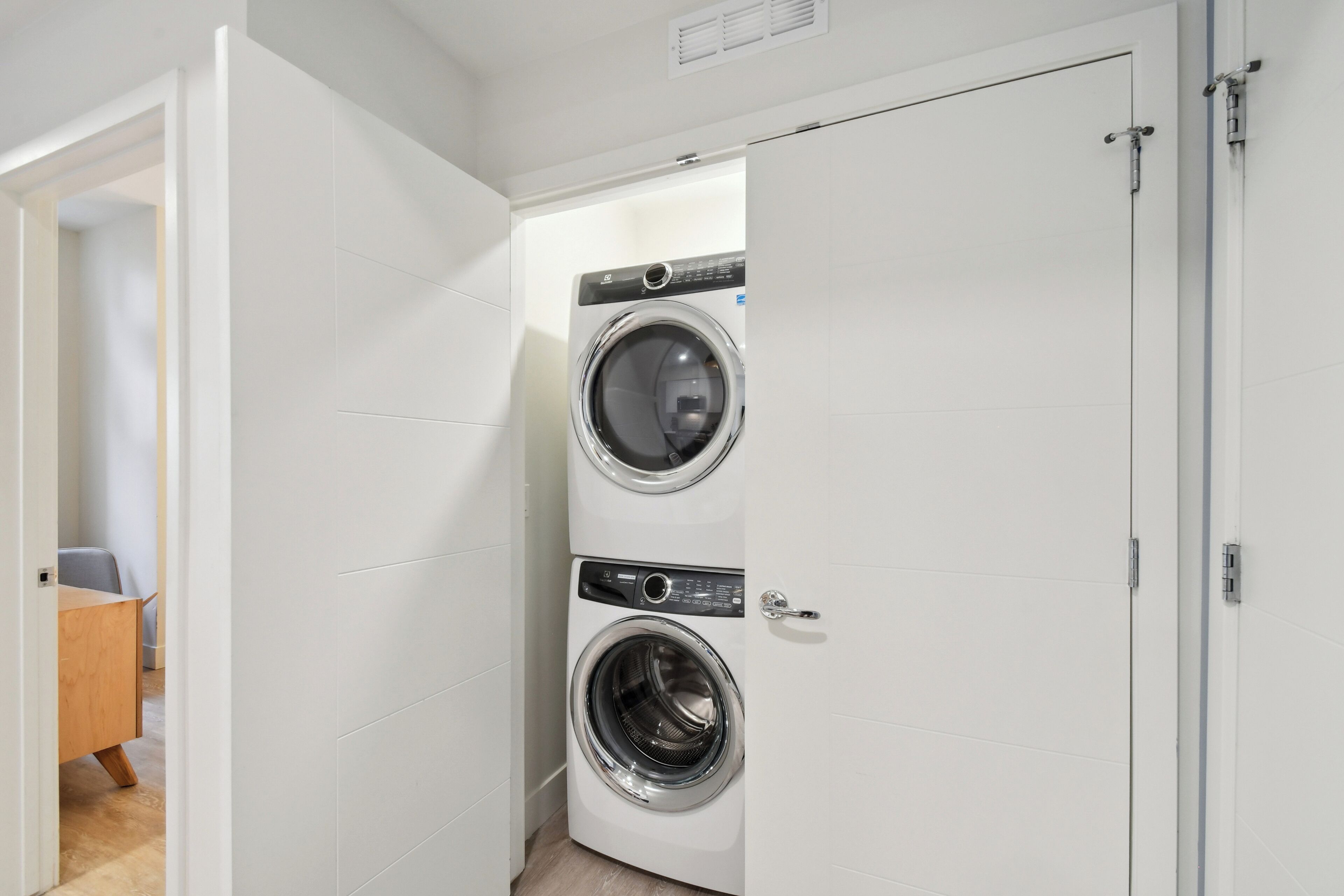A laundry room with two stacked front-loading washing machines, a wooden desk, and a door leading to another room.