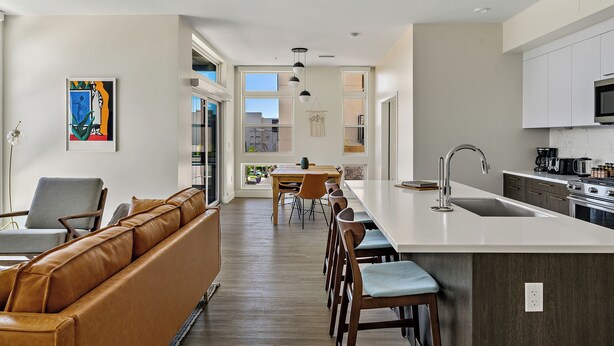 A modern kitchen with a central island, a dining area with a sofa and chairs, and a large window providing natural light.