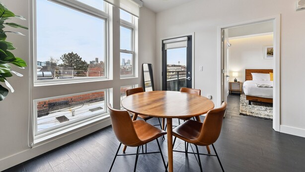Une chambre moderne avec une table ronde en bois et des fauteuils en cuir, de grandes fenêtres donnant sur la ville, un lit avec une couette blanche et un luminaire suspendu.