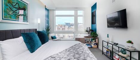 A modern bedroom with a large bed, a city view, a mounted TV, and a bookshelf.