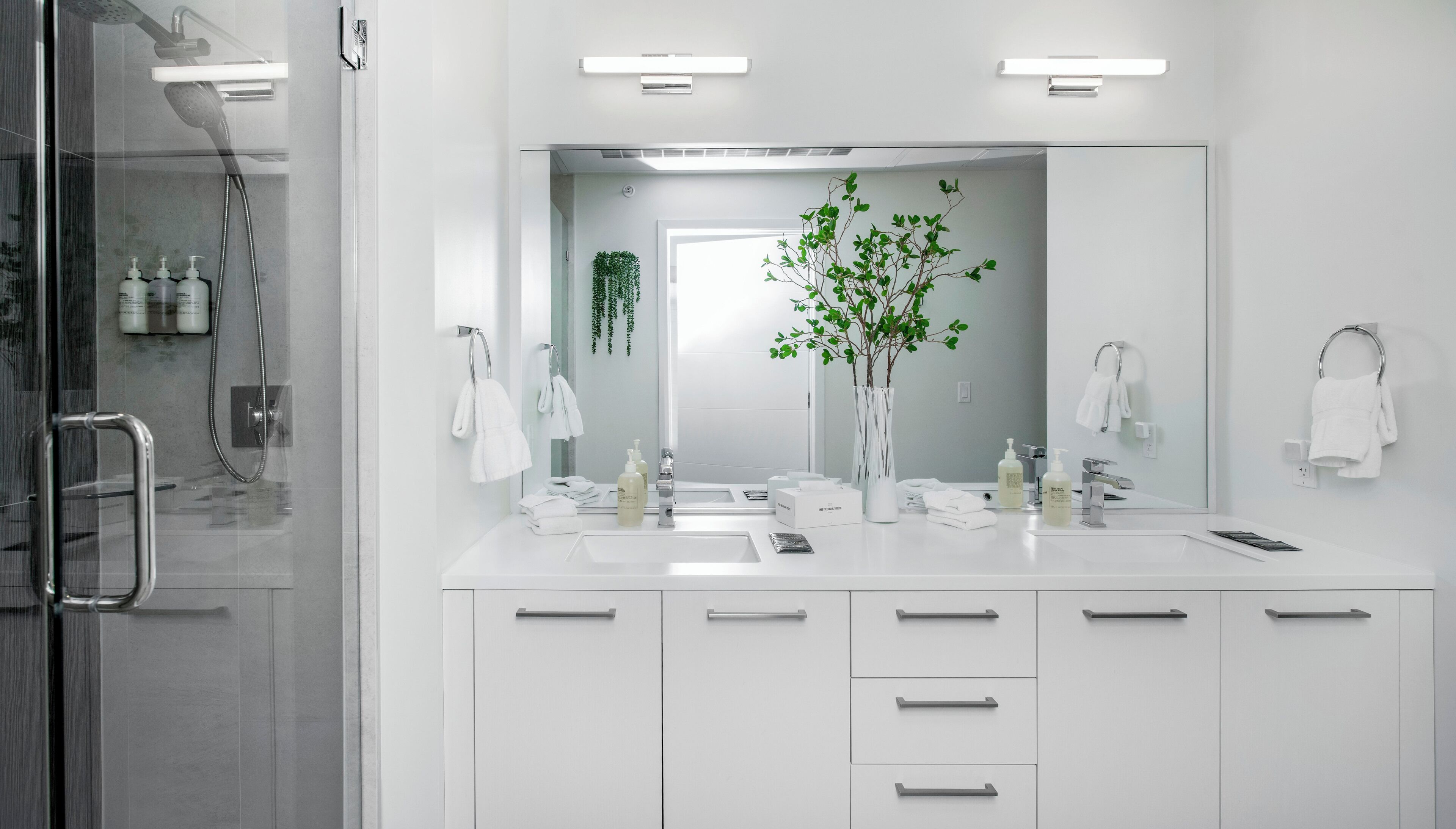 Ein modernes Badezimmer mit einem großen Spiegel, zwei Waschbecken, einer Dusche und einer Pflanze.