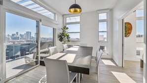 A modern dining area with a large table, white chairs, and a city view through large windows.