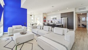 A modern living room with a large sectional sofa, a dining area, and a kitchen with dark cabinets and stainless steel appliances.