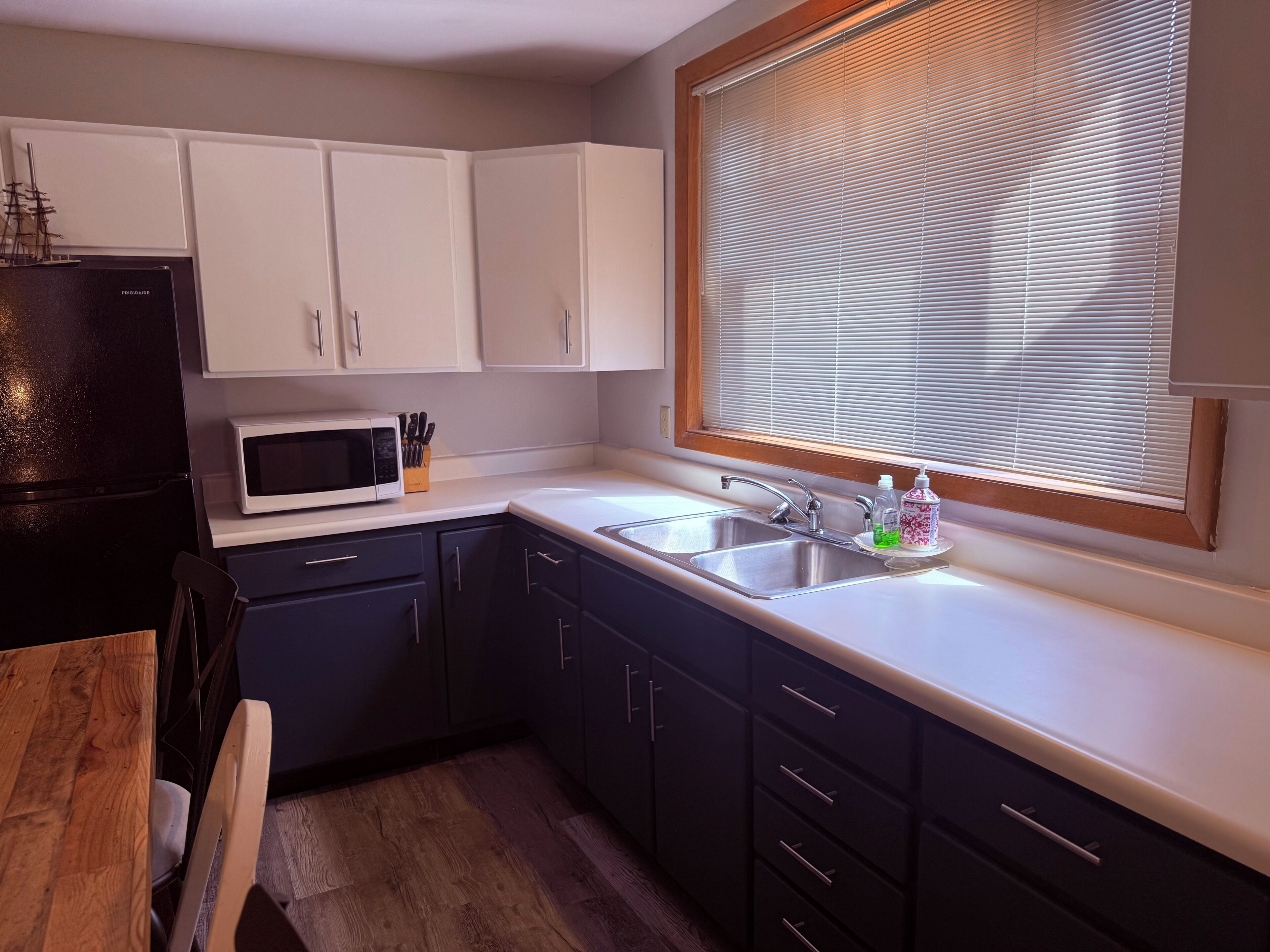 Una cocina con refrigerador negro, microondas blanco y un fregadero con ventana arriba.