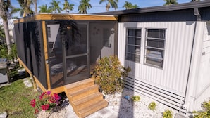 Una casa móvil con terraza de madera, puerta corrediza de vidrio y un pequeño jardín con flores y arbustos.