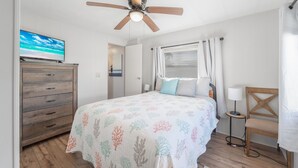 Un dormitorio con cama, ventilador de techo, televisor, cómoda, ventana con cortinas y mesita de noche con lámpara.