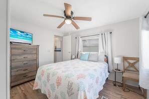 A bedroom with a bed, a ceiling fan, a TV, a dresser, a window with curtains, and a nightstand with a lamp. - Tiny Treasure (Fort Myers)