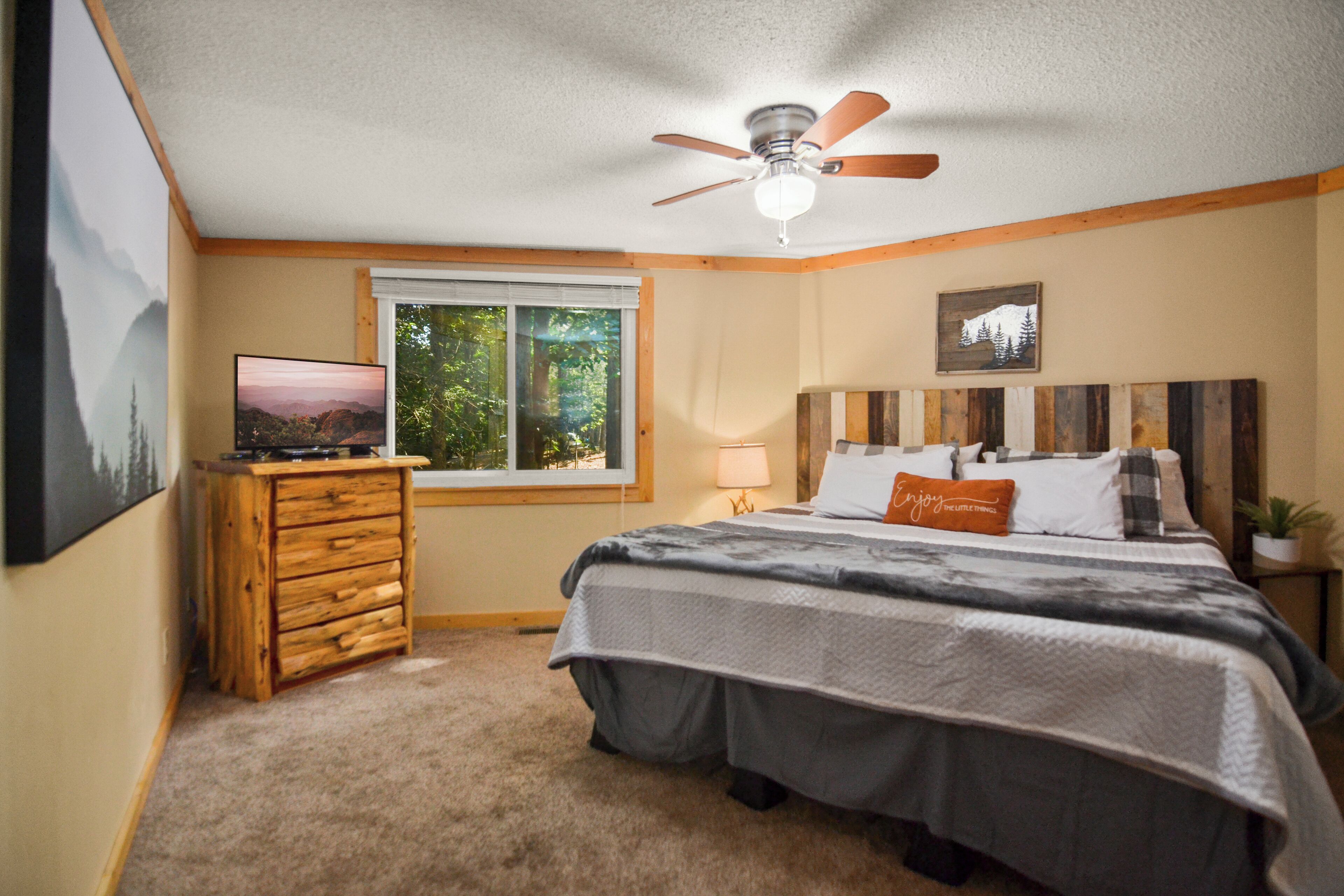 A bedroom with a bed, a ceiling fan, a TV on a wooden dresser, a window with a view of trees, and a wall-mounted painting.