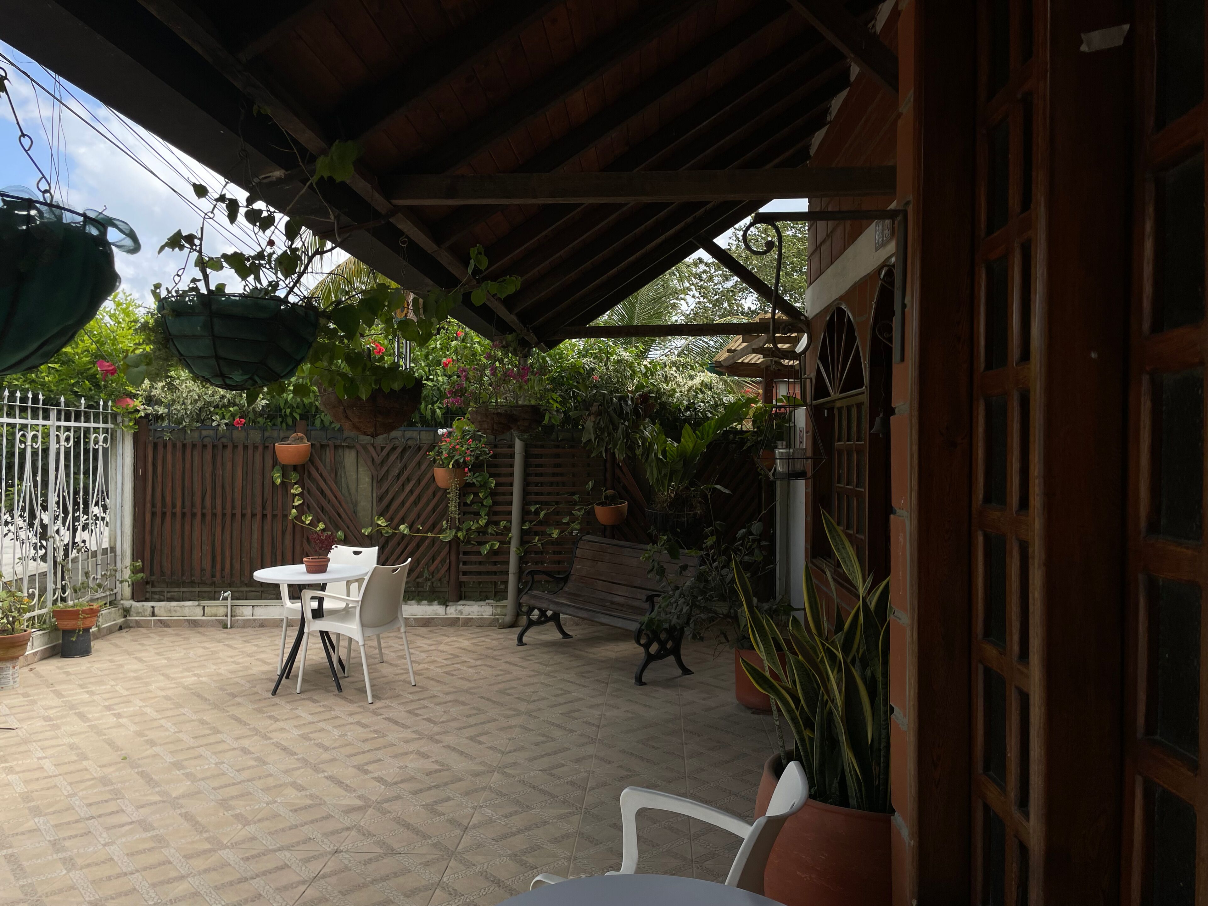 An outdoor patio area with a covered seating area, a table, and chairs. There are potted plants and a wooden fence in the background.