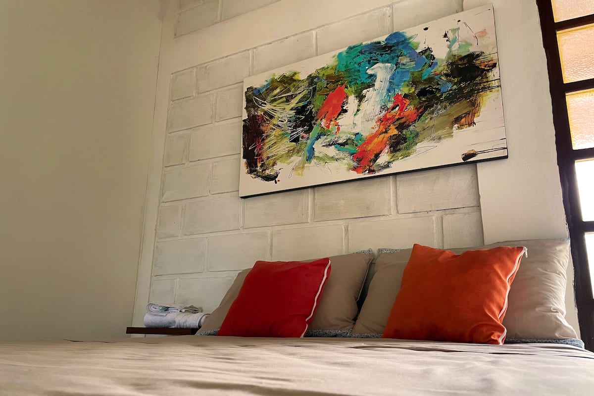 Une chambre à coucher avec un lit, des oreillers et un tableau abstrait aux couleurs vives accroché au mur.
