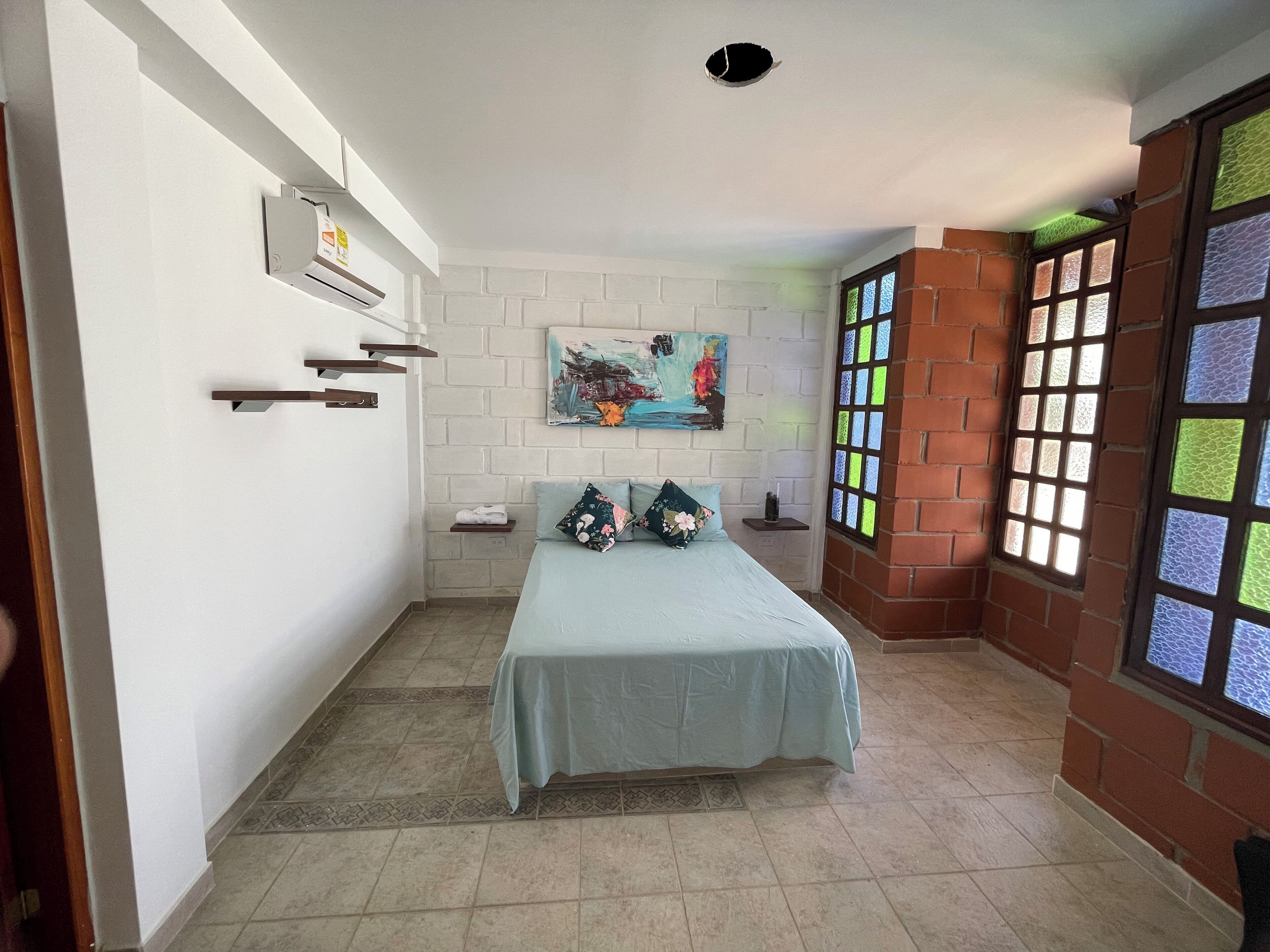 A bedroom with a bed, a painting on the wall, a window with stained glass, and a brick wall.