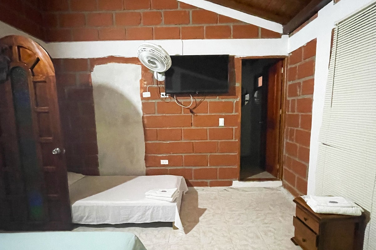 Une petite pièce peu éclairée, avec un mur en briques, un plafond en bois, un lit avec un matelas blanc, une couverture verte, une petite table de chevet en bois et un ventilateur fixé au mur.