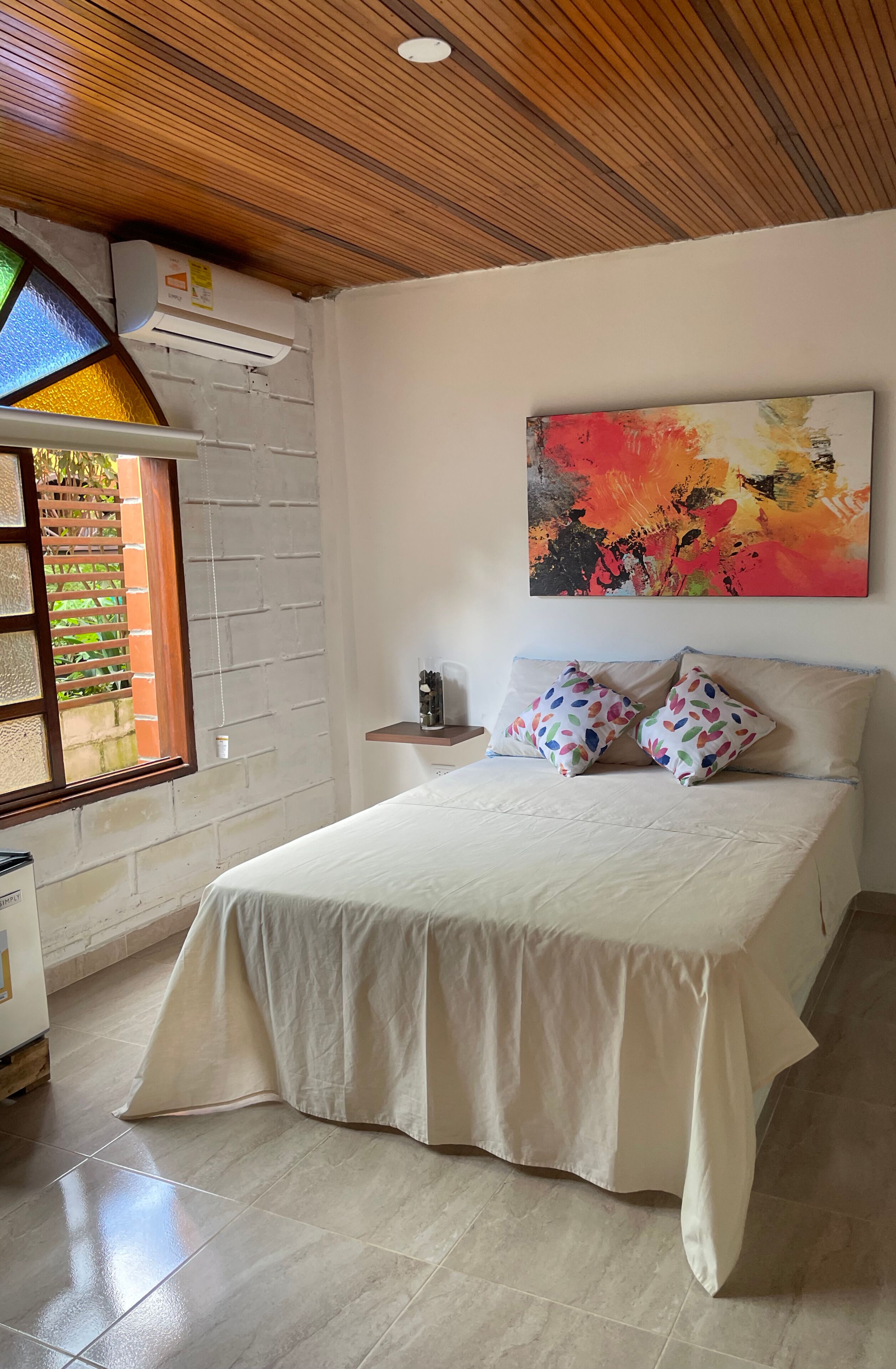 A bedroom with a bed, a window, a painting, and a wall-mounted air conditioner.