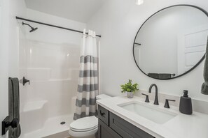 A bathroom with a shower, toilet, sink, and a round mirror. - Gorgeous New Home, Sleeps 10, Steps Away from Lake Michigan Beaches (Muskegon)