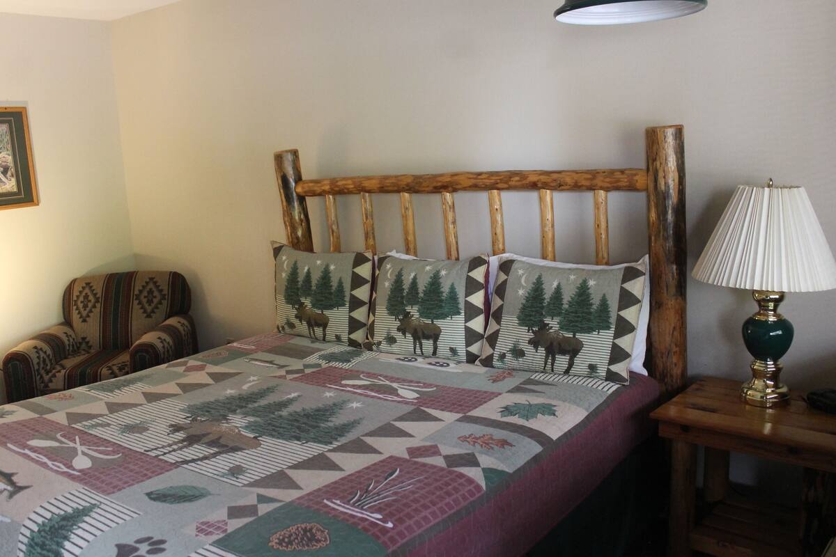 A bed with a geometric patterned bedspread, a wooden headboard, two matching pillows, a wooden nightstand with a lamp, and a chair with a patterned cushion.