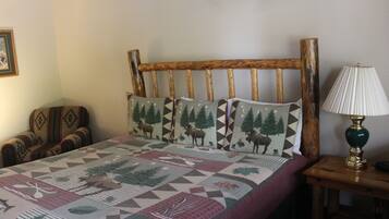 A bed with a geometric patterned bedspread, a wooden headboard, two matching pillows, a wooden nightstand with a lamp, and a chair with a patterned cushion.
