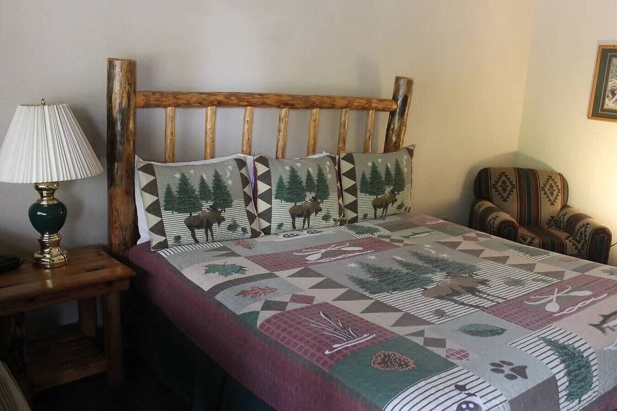 A bed with a wooden headboard, two matching pillows, a patterned bedspread, a lamp on a bedside table, and a chair with a patterned cushion.