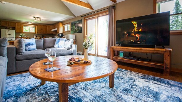 A cozy living room with a wooden coffee table, a grey sofa, and a flat-screen TV displaying a fireplace. - Trailside Lodge- Trail Access w/Game Table-New AC (Pittsburg)