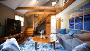 A cozy living room with a wooden staircase, a flat-screen TV, a round coffee table, and a grey sofa. - Trailside Lodge- Trail Access w/Game Table-New AC (Pittsburg)
