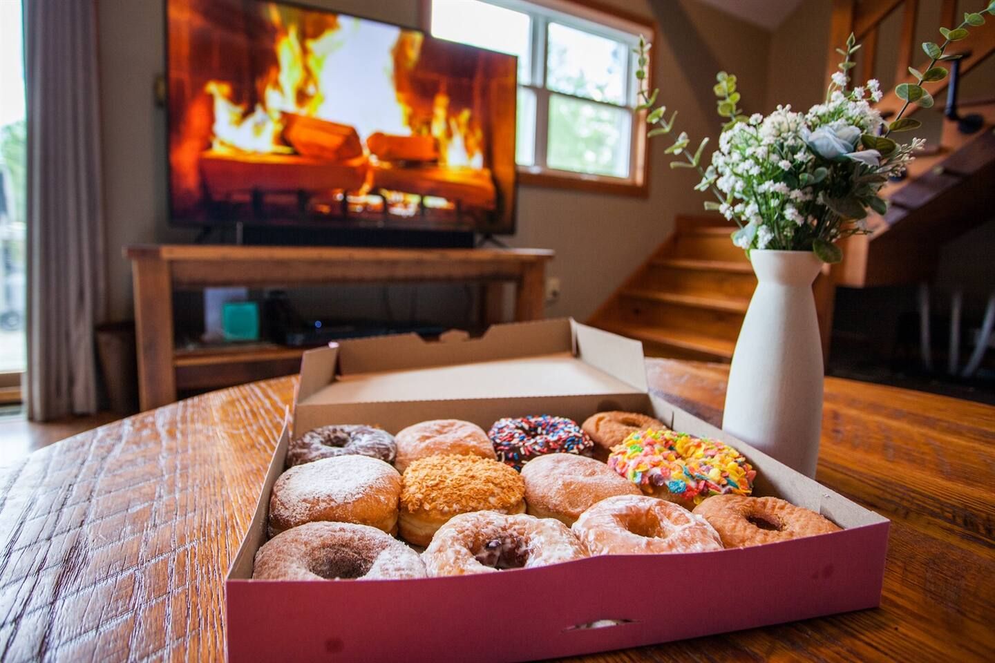 Een doos met diverse donuts op een houten tafel, met een televisie waarop een brand te zien is en een bloevas ernaast.