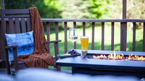A wooden deck with a table, a chair, a blanket, a glass of white wine, a glass of orange juice, and a fire pit. - Trailside Lodge- Trail Access w/Game Table-New AC (Pittsburg)