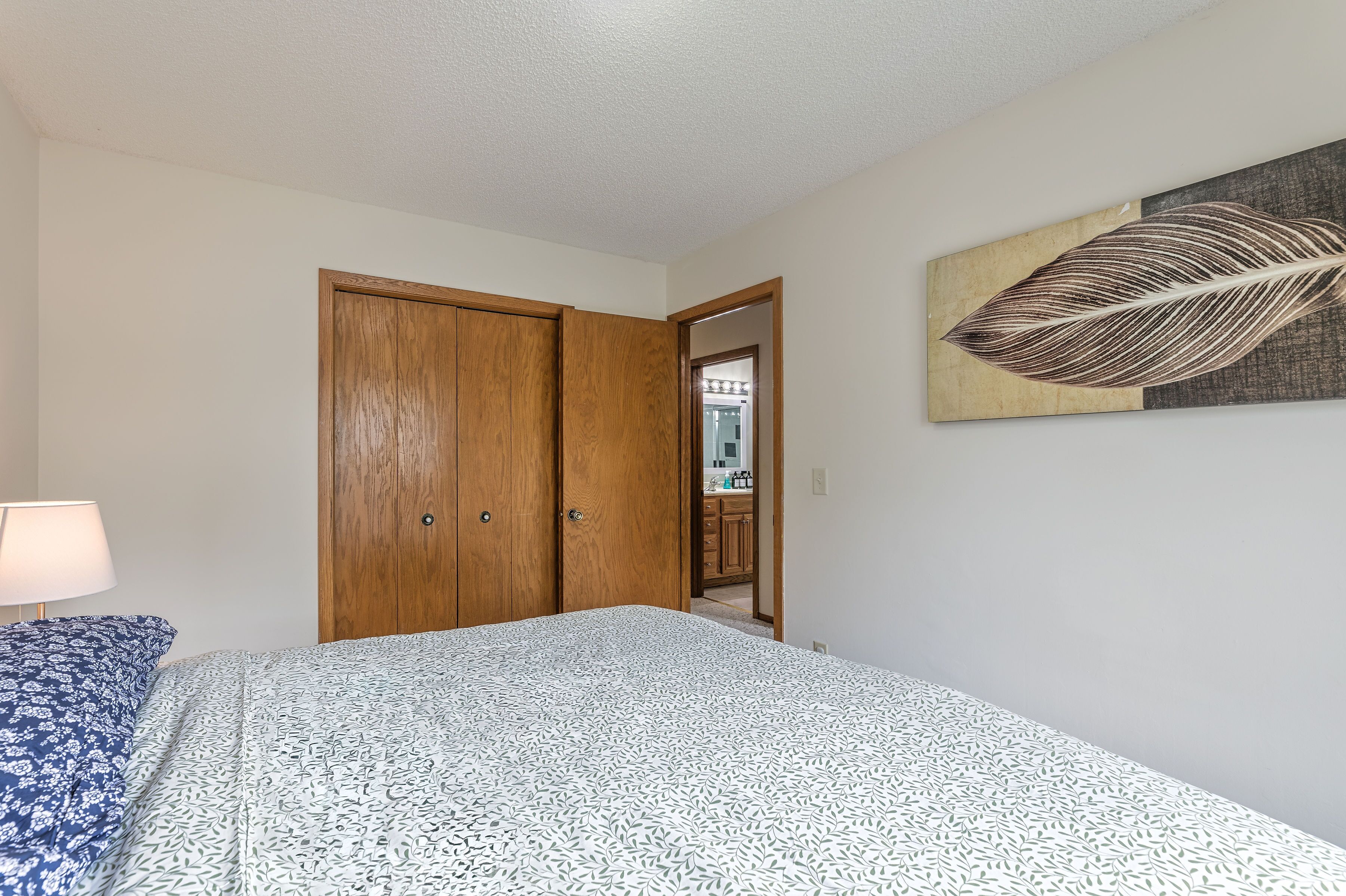 Une chambre à coucher avec un lit, une armoire en bois, une œuvre d’art accrochée au mur, et une salle de bain visible à travers une porte ouverte.
