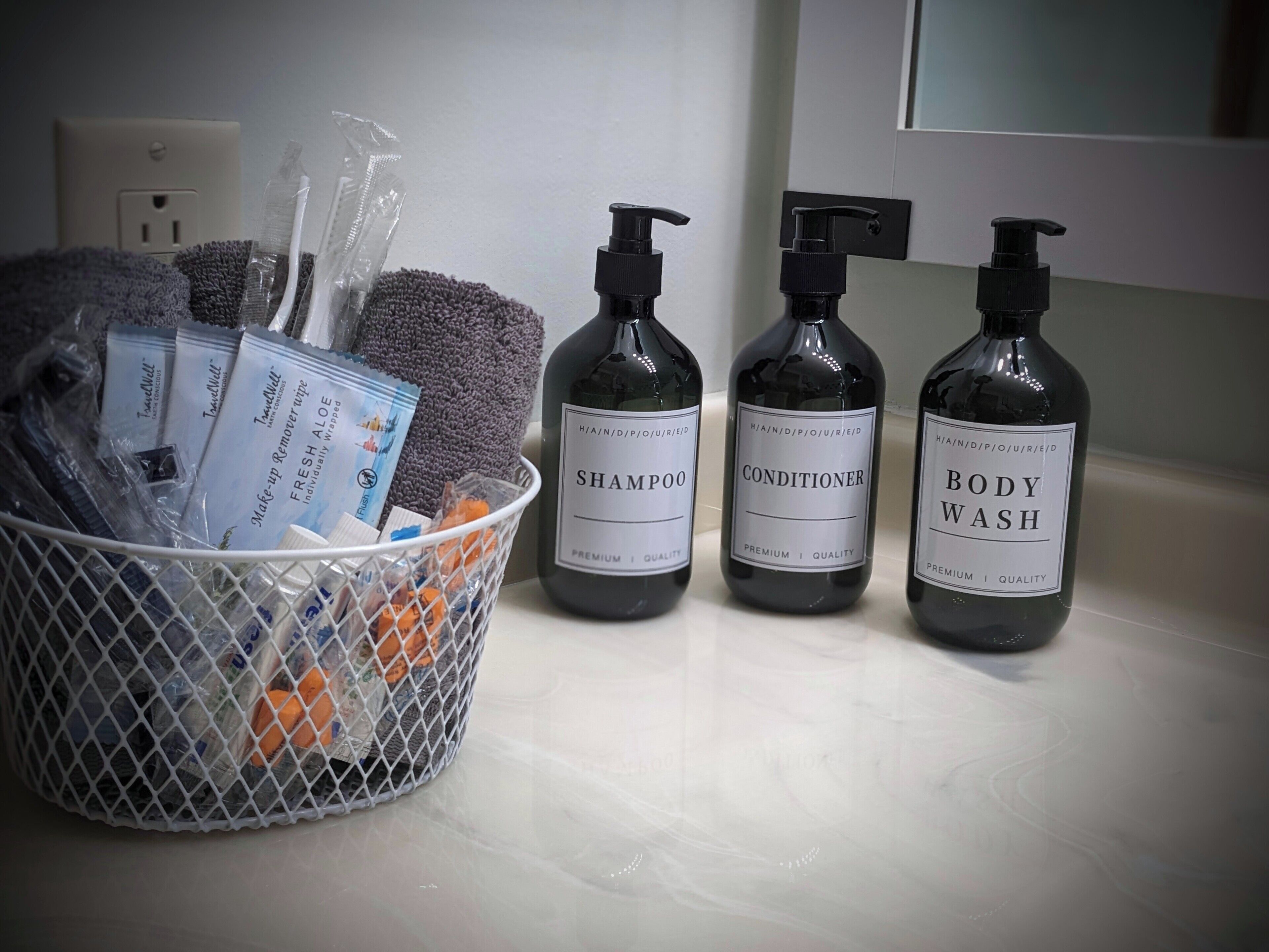 A bathroom with a white sink, three bottles of toiletries labeled shampoo, conditioner, and body wash, and a basket containing various items including a towel and toiletries.