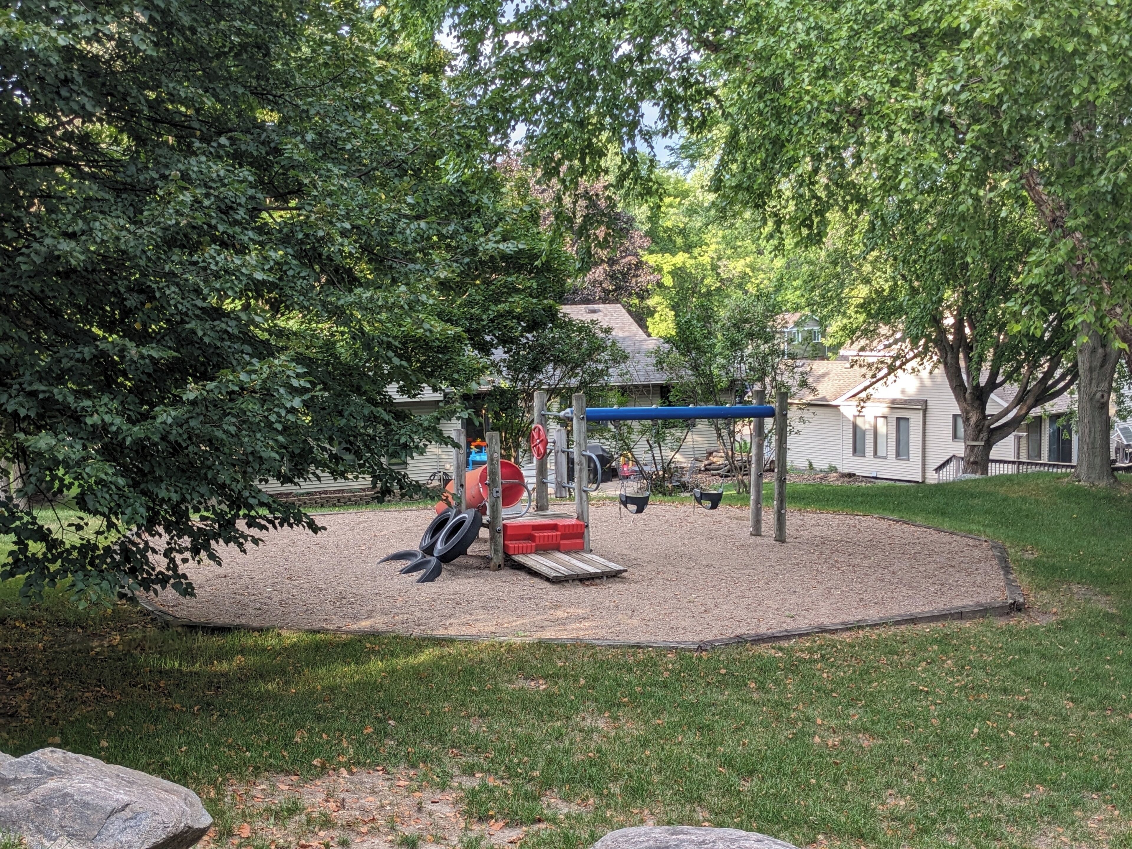 Un parque infantil con columpios y un tobogán, rodeado de árboles y una casa al fondo.