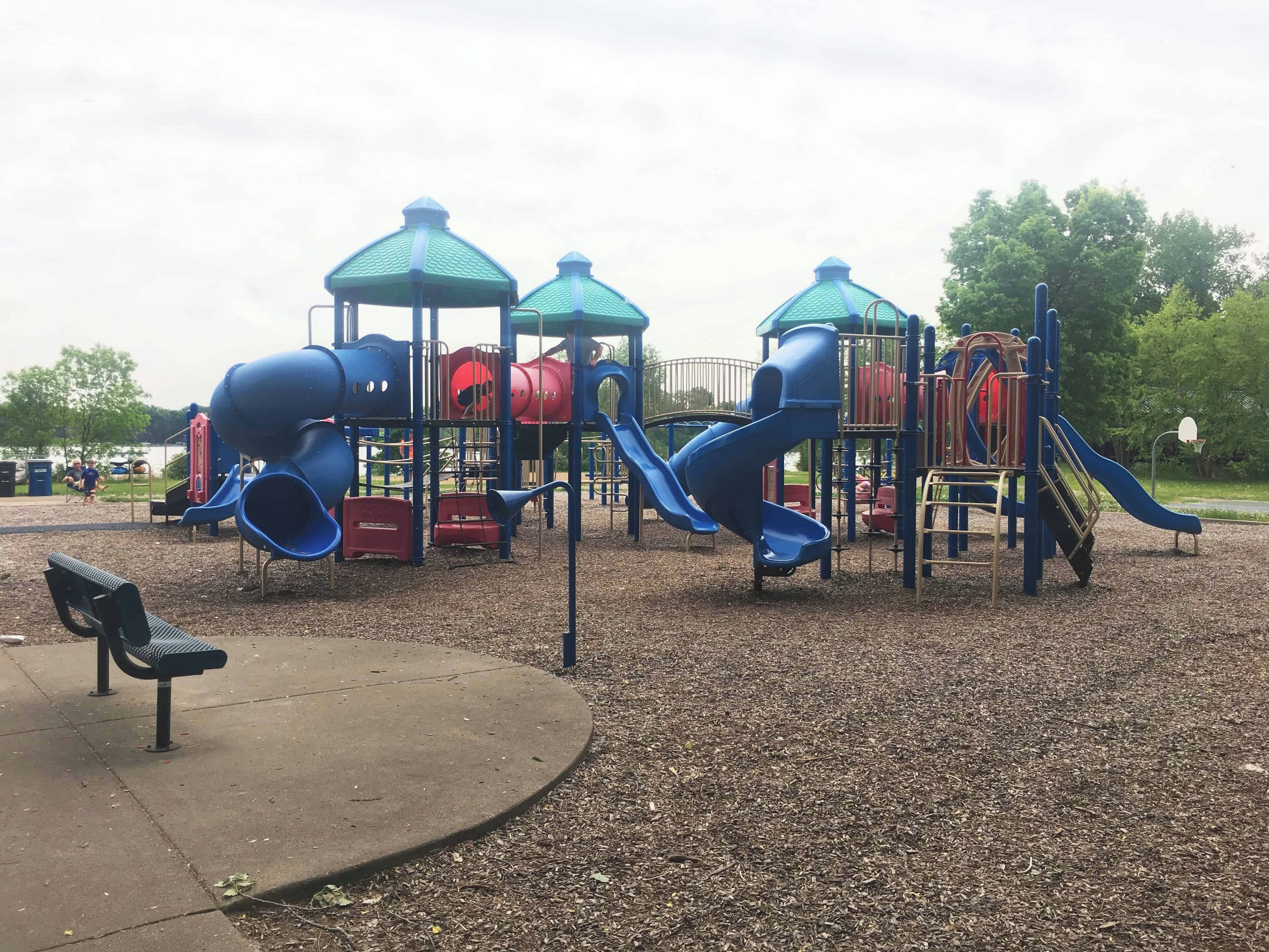 A playground with multiple slides and climbing structures, surrounded by a gravel area and a bench.