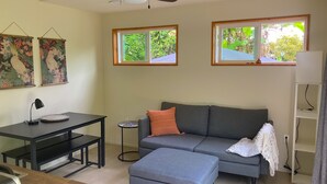 Un salon avec un canapé gris, une petite table ronde, un bureau avec une lampe et deux fenêtres donnant sur un espace verdoyant.