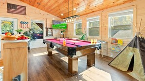 Ein Billardtisch in einem Raum mit Holzwänden und -decke, ein Fenster mit Blick auf Bäume und ein kleiner Tisch mit einem Cupcake und Obst.