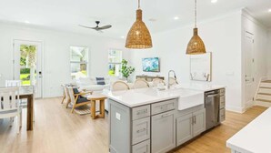 A modern kitchen with grey cabinets, a white countertop, and a central island. There are wicker pendant lights, a ceiling fan, and a view of the outdoors through large windows. - Vitamin Sea by Stay on 30a (Inlet Beach)