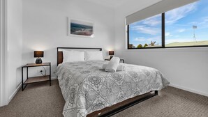 Une chambre à coucher moderne avec un grand lit, deux tables de chevet avec des lampes, un tableau accroché au mur et une grande fenêtre offrant une vue sur le ciel et le paysage lointain.