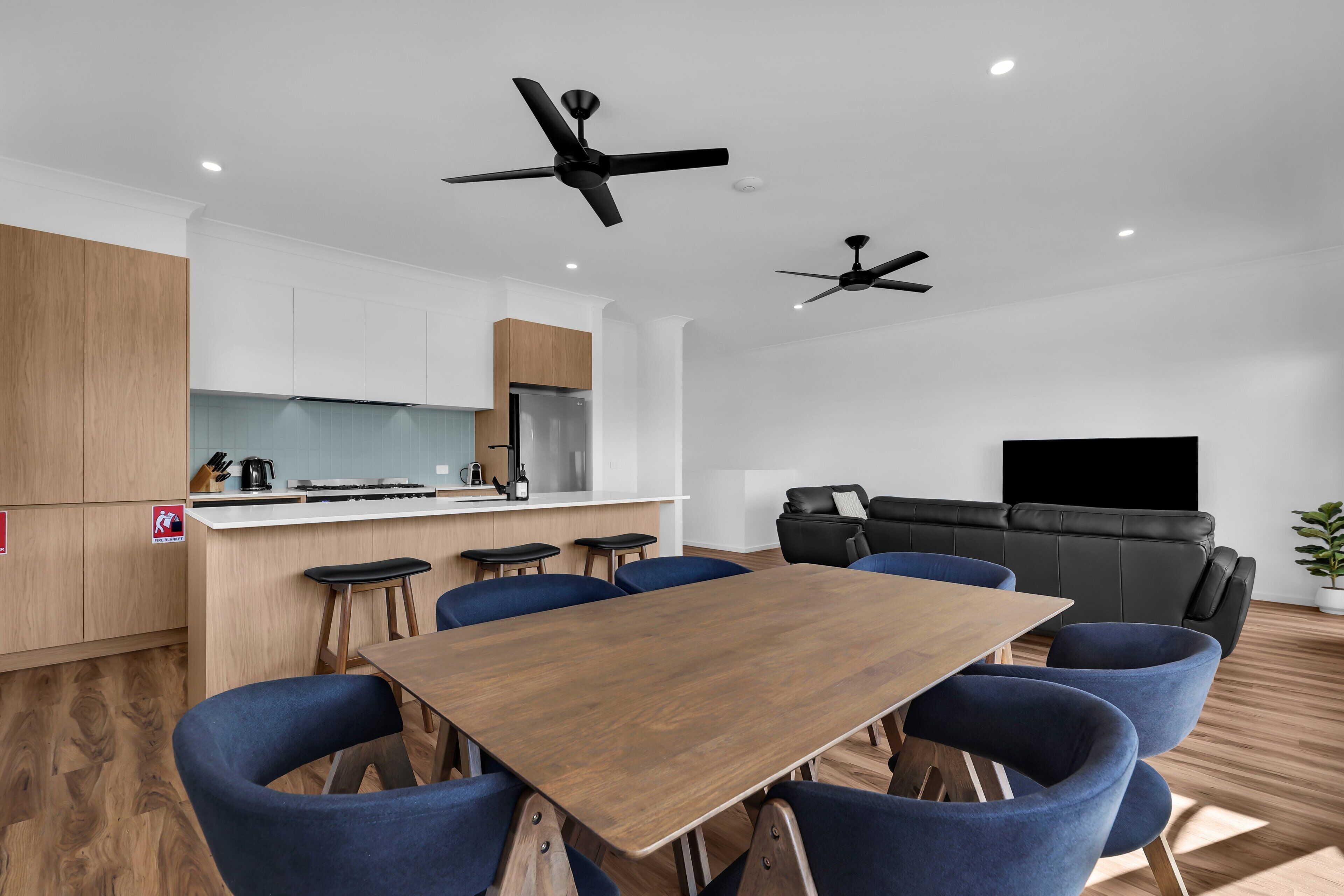 A modern kitchen with wooden cabinets, a bar area, and a dining space with blue chairs. There are ceiling fans and a flat-screen TV in the living area.
