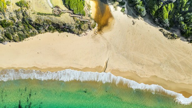 Une vue aérienne d’une plage aux eaux turquoise et limpides, avec un rivage sablonneux et une promenade en bois menant à une construction.