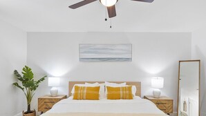 Un dormitorio con cama, dos mesitas de noche con lámparas, un ventilador de techo, un espejo grande y una planta en maceta.