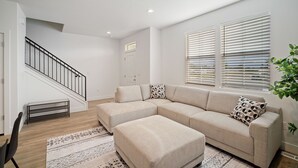 A modern living room with a sectional sofa, a small ottoman, a staircase, and a large window with blinds.